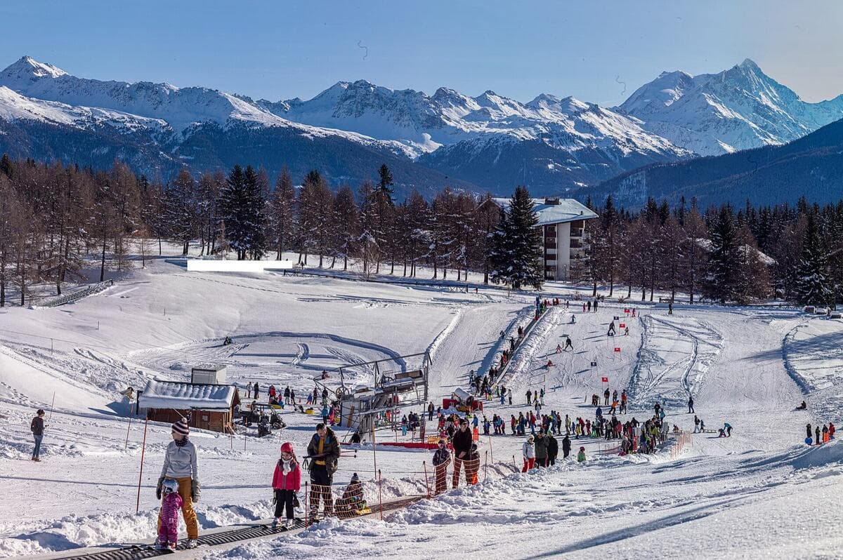 A parent and small child on the magic carpet at a busy beginner area below snowy peaks