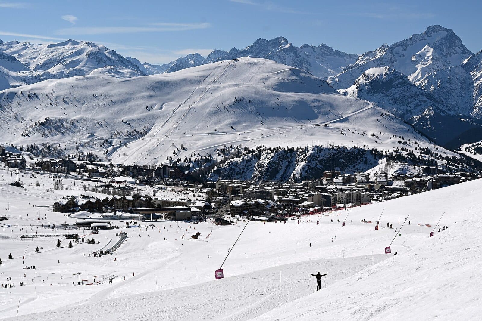 Skiers on broad sunny pistes above Alpe d'Huez, with high snowy peaks beyond the resort