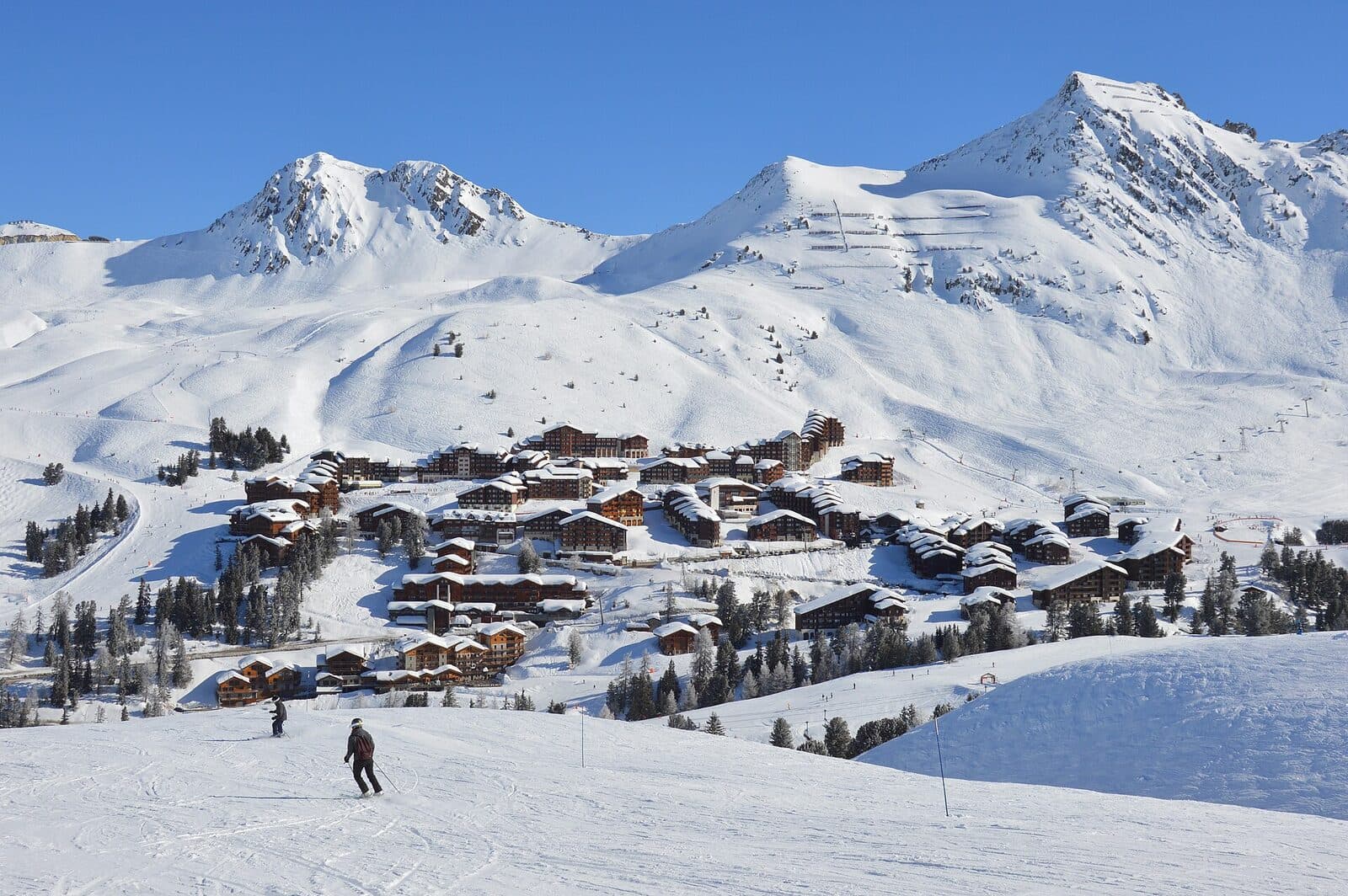 The chalet-style buildings of Belle Plagne below snowy peaks, with skiers on the piste in front