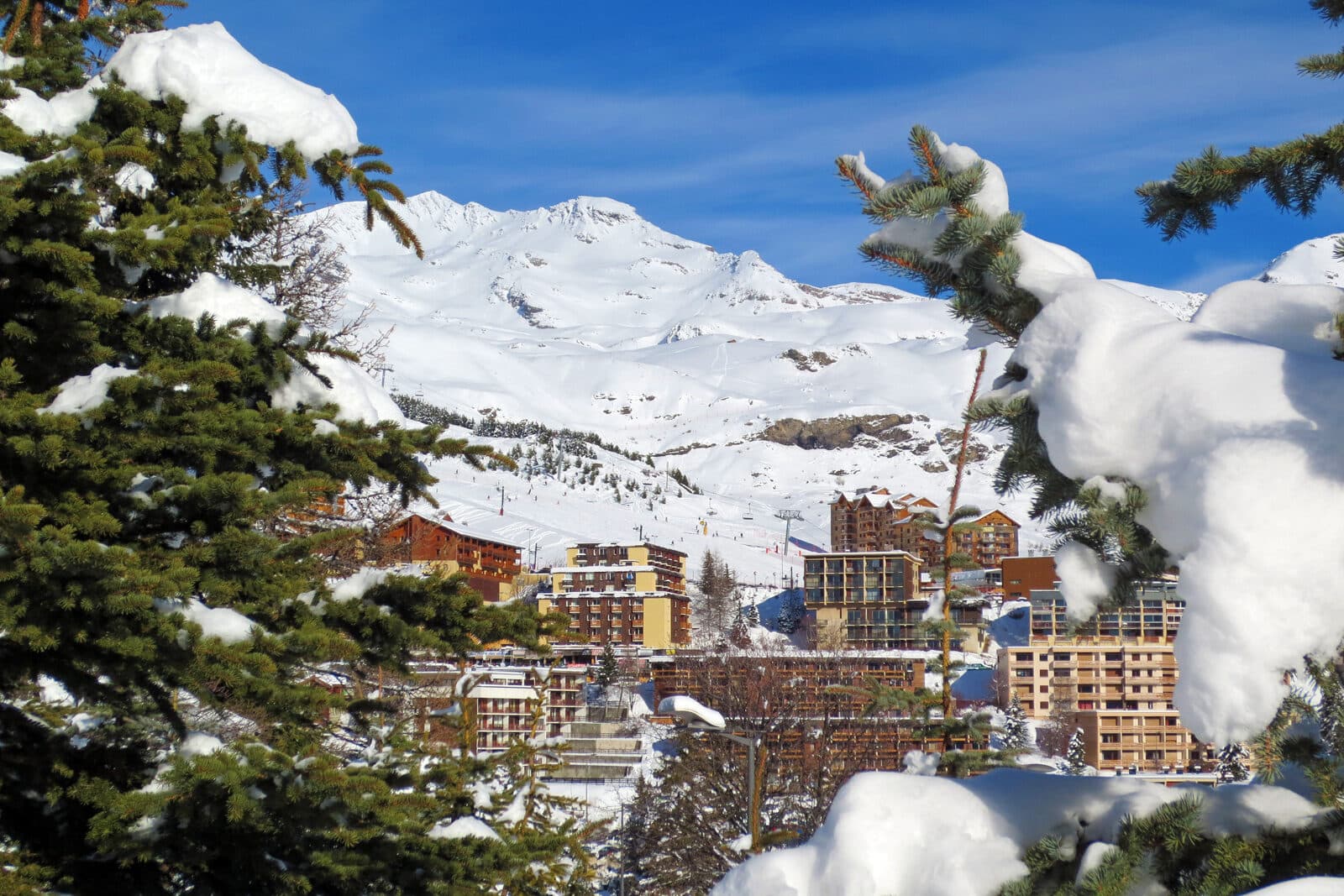 The apartment buildings of Orcières Merlette 1850 seen through snow-laden firs, with sunny pistes and peaks behind